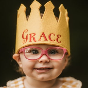 Puede incluir: Un ni&ntilde;o sonriente con una corona de fieltro amarillo con el nombre "GRACE" bordado en rojo. El ni&ntilde;o tambi&eacute;n lleva gafas con montura rosa. El fondo es un verde oscuro borroso.
