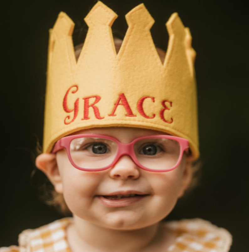 Puede incluir: Un ni&ntilde;o sonriente con una corona de fieltro amarillo con el nombre "GRACE" bordado en rojo. El ni&ntilde;o tambi&eacute;n lleva gafas con montura rosa. El fondo es un verde oscuro borroso.