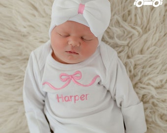 May include: A white baby gown with long sleeves and a pink bow embroidered on the front. The gown has the name "Harper" embroidered below the bow. The baby is wearing a white knit hat with a pink bow on top.