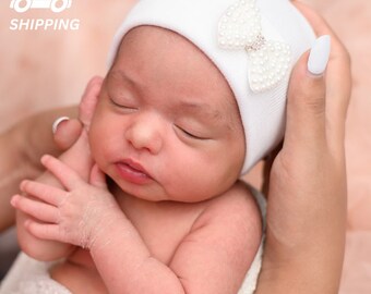 May include: A white baby hat with a pearl bow. The hat is made of soft, stretchy fabric and is perfect for newborns.