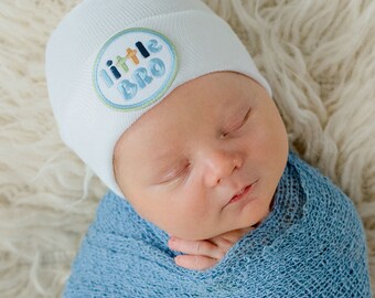 May include: A white baby hat with a blue and green embroidered patch that says "little bro". The hat is on a baby's head, who is sleeping and wrapped in a blue blanket.