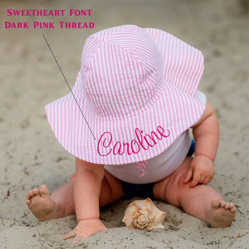May include: A pink and white striped sun hat with the name "Caroline" embroidered in dark pink thread. The hat has a wide brim and is made of seersucker fabric. The text "Sweetheart Font Dark Pink Thread" is also visible.