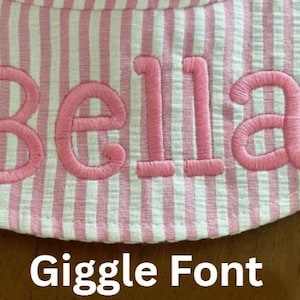 May include: Close-up of a pink and white striped fabric with the name "Bella" embroidered in pink thread. The text "Giggle Font" is displayed below the name. The fabric appears to be part of a hat or similar item.