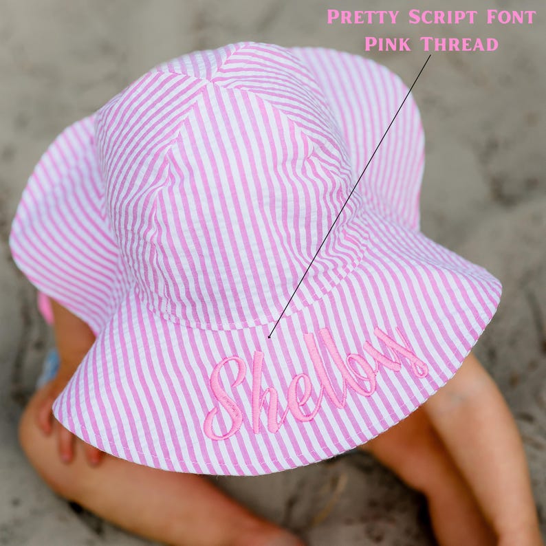 May include: A pink and white striped sun hat with the name "Shelley" embroidered in a pretty script font with pink thread. The hat is designed with a wide brim and is made of a textured fabric.