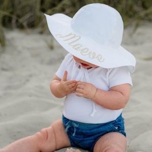 May include: A white sun hat with a wide brim and a gold embroidered name "Maeve" on the front. The hat is worn by a child sitting on a sandy beach.