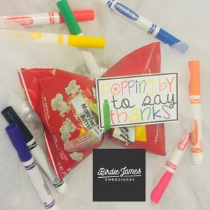 May include: A red and white popcorn bag with a green ribbon and a white card with the text "popping by to say thanks" in colorful letters. The bag is surrounded by colorful markers.
