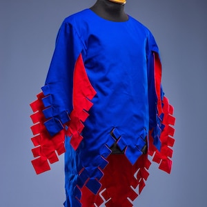 May include: A blue tunic with red detailing, presented on a mannequin. The garment has a round neckline and long sleeves. Red, square cutouts adorn the sleeves and hem. The Steel Mastery logo is visible at the bottom of the image.