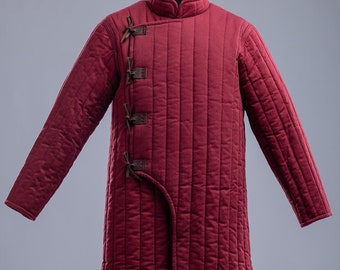Puede incluir: Una chaqueta larga acolchada de color burdeos con cuello alto y una fila de correas de cuero en la parte delantera. La chaqueta tiene un dobladillo ligeramente acampanado y mangas largas.