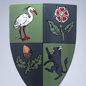 May include: A green and gray shield with four different images. The top left image is a white heron with a red beak. The top right image is a red rose with green leaves. The bottom left image is an acorn with green leaves. The bottom right image is a black bull with green horns.