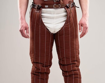 May include: Brown quilted leg armor with a leather belt and buckles. The armor is worn over white undergarments.