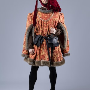 Può includere: Una persona in costume rinascimentale. L'outfit presenta una tunica fantasia rossa e oro con finiture in pelliccia, un cappuccio rosso abbinato e leggings neri. Una borsa in pelle nera è appesa a una cintura.