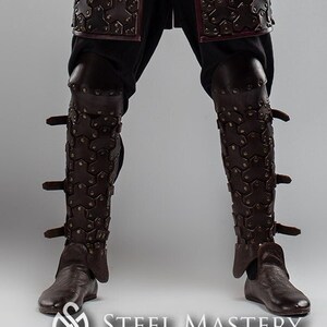 May include: A person wearing brown leather armor with a crossbow strapped to their shoulder. The armor has a studded design and is worn over a black shirt and pants. The person is standing with their right arm raised, holding the crossbow.