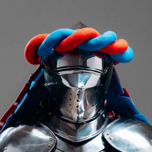 Op de afbeelding: Een ridder in volledige bepantsering met een blauw-rode helm en een blauw-rode cape. Het harnas is gepolijst en glanzend. De ridder draagt een helm met een vizier. De helm is versierd met een blauw-rode tulband. De ridder staat voor een grijze achtergrond. "STEEL MASTERY" staat onderaan de afbeelding.