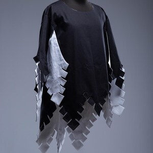 May include: A black tunic with long sleeves and a distinctive design. It has a white panel on the side and a decorative, layered hemline. The hemline is made of alternating black and white geometric shapes.