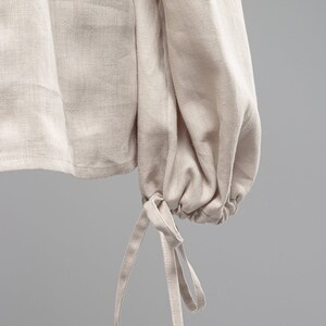 May include: A close-up of a beige linen shirt with long sleeves and a gathered cuff detail. The cuff has two fabric ties that can be tied into a bow.