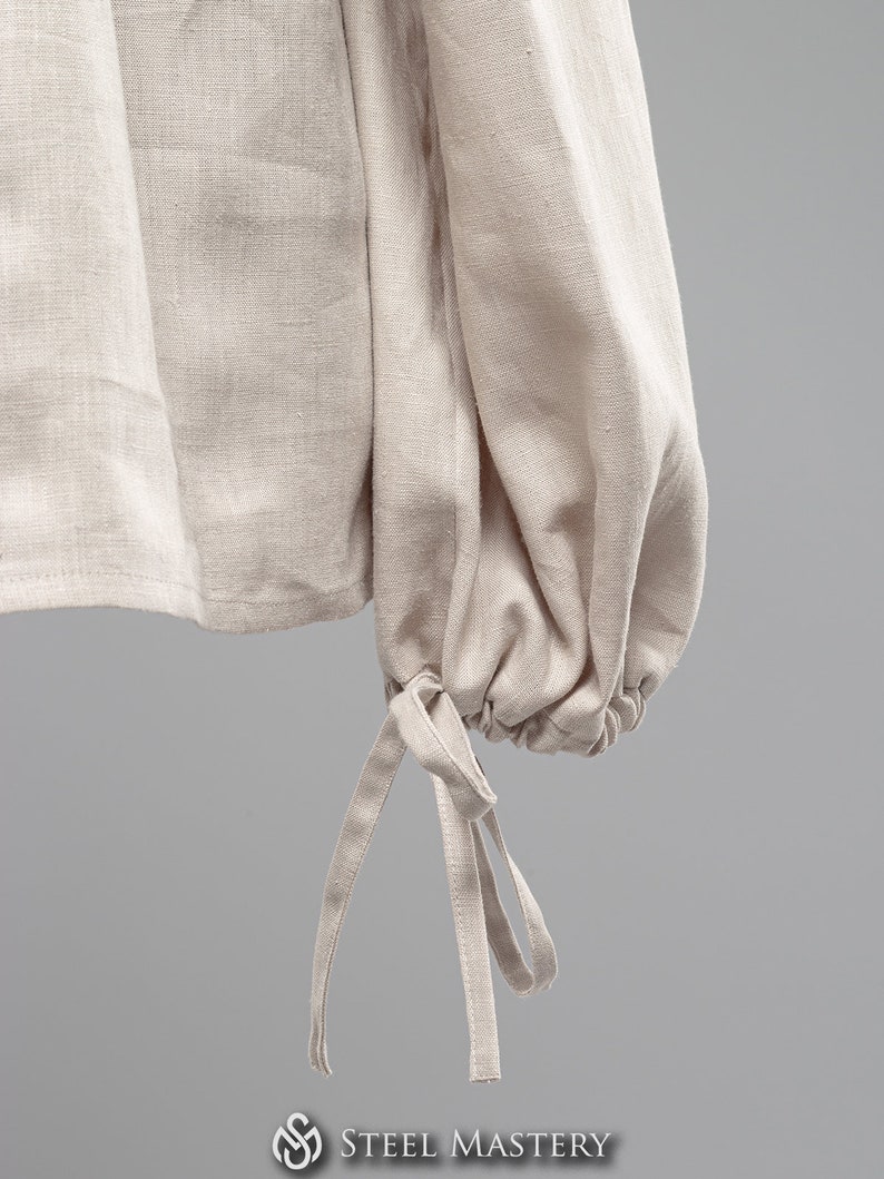 May include: A close-up of a beige linen shirt with long sleeves and a gathered cuff detail. The cuff has two fabric ties that can be tied into a bow.