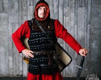 以下が含まれることがあります： 赤いチュニックと、茶色のフードが付いた黒いスタッズベストを着た人物。その人物は剣と盾を持っています。この衣装は中世の鎧を彷彿とさせます。