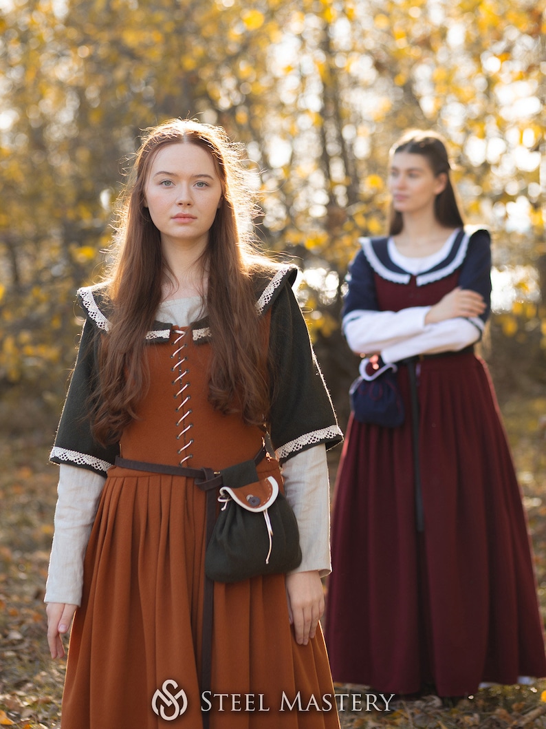 May include: Two women wearing medieval-style dresses stand in a forest. The woman in the foreground is wearing a brown dress with a green bodice and a white lace trim. She is also wearing a brown leather belt and a small green pouch. The woman in the background is wearing a burgundy dress with a white collar and a blue sash. She is standing with her arms crossed.