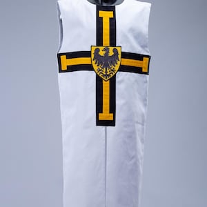 May include: White tunic with a black and yellow cross design. The tunic features a shield with an eagle emblem at the center of the cross. The garment is displayed on a black mannequin. The Steel Mastery logo is at the bottom.