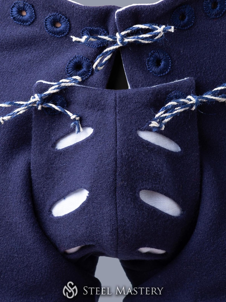 Op de afbeelding: Close-up van de achterkant van een paar donkerblauwe middeleeuwse broeken met witte veters en witte oogjes. De broek heeft een uniek ontwerp met uitsparingen op de rug.
