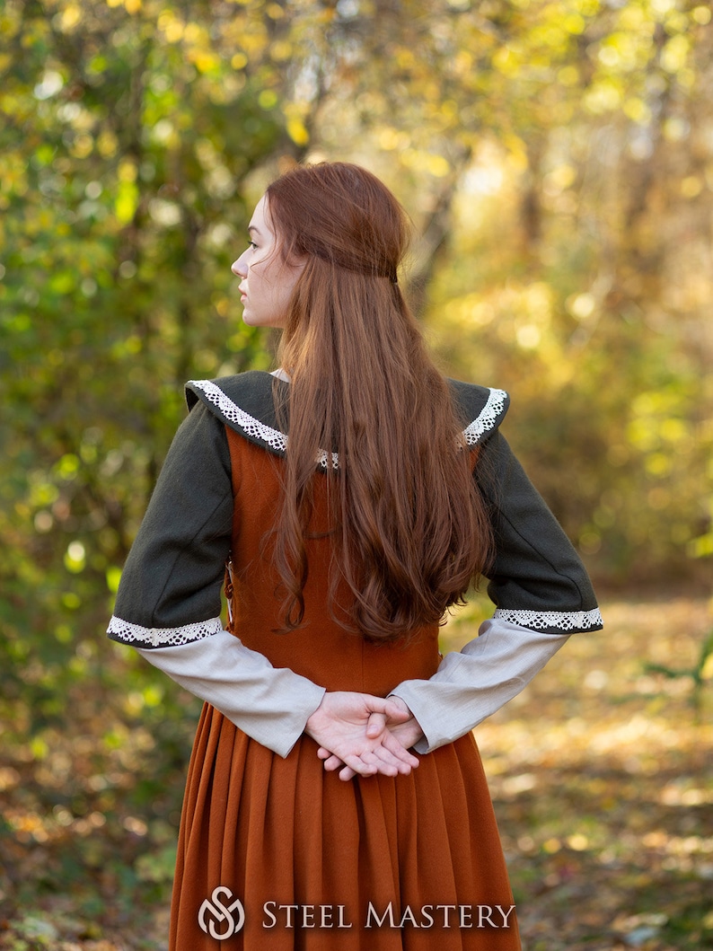 May include: A woman wearing a long, rust-colored dress with a green and white lace trim. She is standing in a forest with fall foliage in the background.