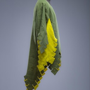 May include: A green tunic with yellow detailing, shown on a mannequin. The tunic has a long, flowing design with a V-neck and decorative cutouts along the sleeves and hem. The yellow fabric is visible on the sleeves and hem. The Steel Mastery logo is at the bottom.