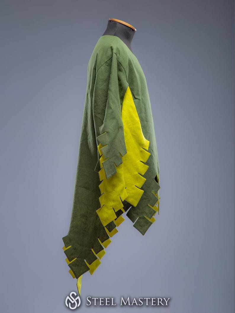 May include: A green tunic with yellow detailing, shown on a mannequin. The tunic has a long, flowing design with a V-neck and decorative cutouts along the sleeves and hem. The yellow fabric is visible on the sleeves and hem. The Steel Mastery logo is at the bottom.