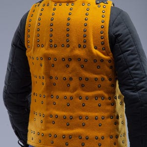 Pode incluir: Colete acolchoado amarelo mostarda com fileiras de rebites. O colete &eacute; usado sobre uma jaqueta acolchoada preta. A parte de tr&aacute;s do colete &eacute; vis&iacute;vel, mostrando o padr&atilde;o de rebites. O logotipo "STEEL MASTERY" est&aacute; na parte inferior.