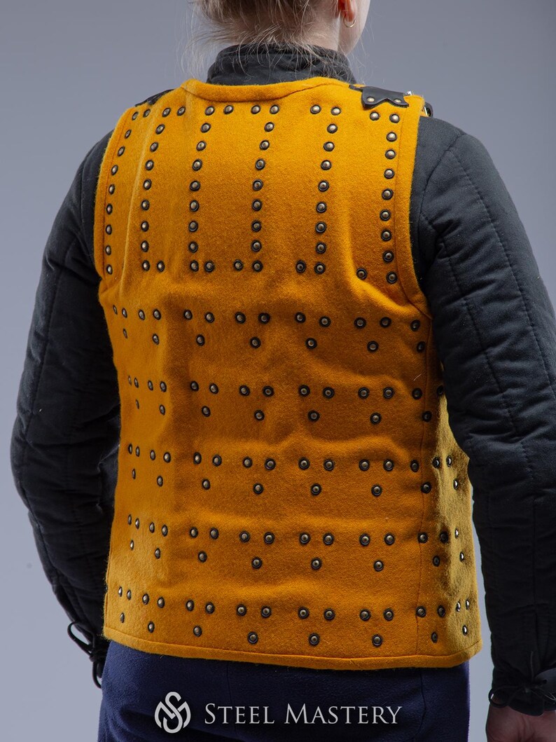 Pode incluir: Colete acolchoado amarelo mostarda com fileiras de rebites. O colete &eacute; usado sobre uma jaqueta acolchoada preta. A parte de tr&aacute;s do colete &eacute; vis&iacute;vel, mostrando o padr&atilde;o de rebites. O logotipo "STEEL MASTERY" est&aacute; na parte inferior.