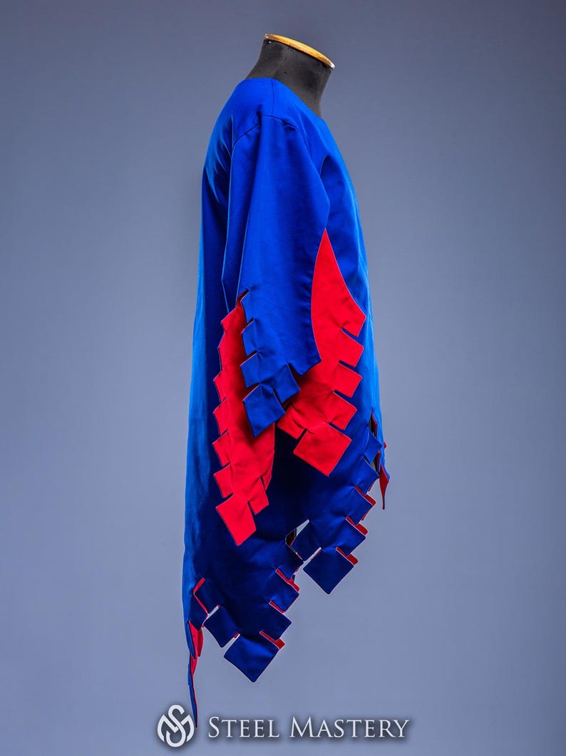 May include: A blue tunic with red detailing, possibly for historical re-enactment. The garment has a long, flowing design with red, rectangular cutouts along the sleeves and hem. The "Steel Mastery" logo is visible at the bottom.