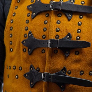 Pode incluir: Close-up de um gambeson medieval cor de mostarda com tiras e fivelas de couro preto. O gambeson &eacute; adornado com in&uacute;meros rebites de lat&atilde;o. O logotipo Steel Mastery &eacute; vis&iacute;vel na parte inferior.