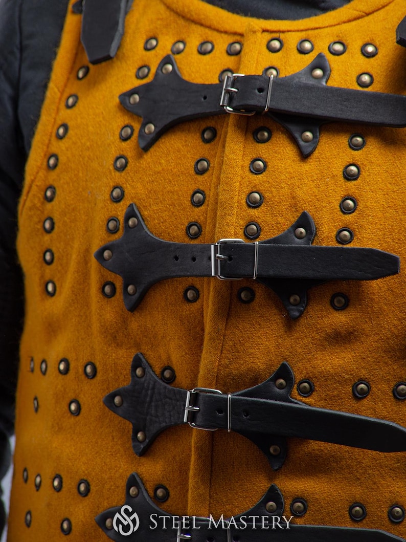 Pode incluir: Close-up de um gambeson medieval cor de mostarda com tiras e fivelas de couro preto. O gambeson &eacute; adornado com in&uacute;meros rebites de lat&atilde;o. O logotipo Steel Mastery &eacute; vis&iacute;vel na parte inferior.