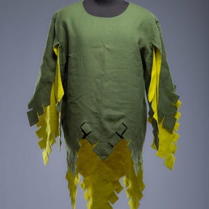 May include: A green tunic with long sleeves and a round neckline. The tunic features yellow accents on the sleeves and hem, with a jagged, decorative edge. The garment is displayed on a mannequin.