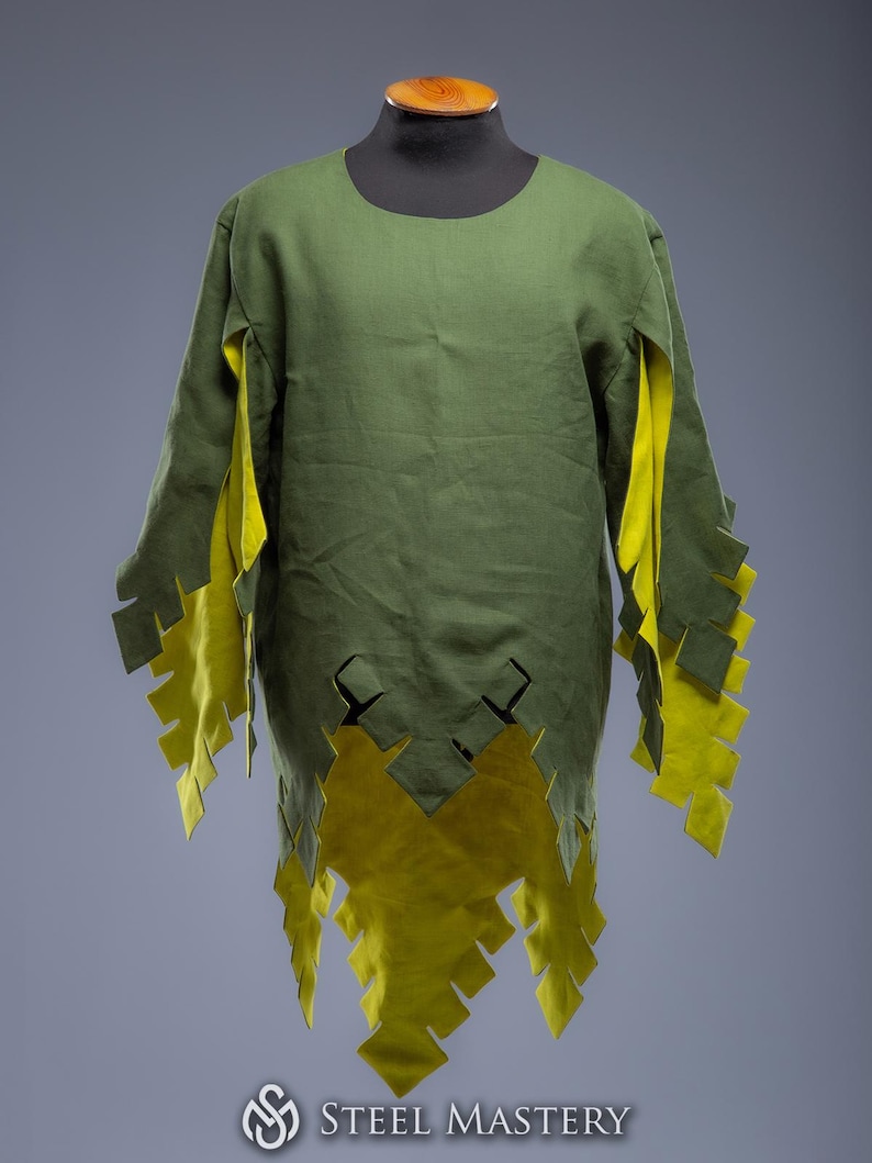 May include: A green tunic with long sleeves and a round neckline. The tunic features yellow accents on the sleeves and hem, with a jagged, decorative edge. The garment is displayed on a mannequin.