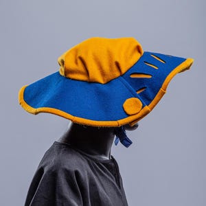K&ouml;nnte beinhalten: Ein blauer und gelber Filzhut mit breiter Krempe und Ausschnitten. Der Hut hat einen runden Knopf an der Seite.