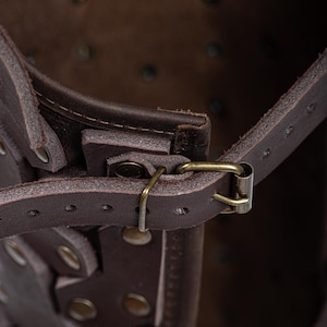 May include: Close-up of a brown leather strap with a metal buckle. The strap is attached to a larger leather piece with rivets. The strap is part of a leather armor piece.