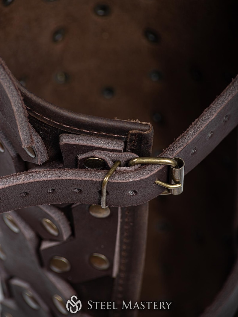 May include: Close-up of a brown leather strap with a metal buckle. The strap is attached to a larger leather piece with rivets. The strap is part of a leather armor piece.