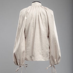 May include: A beige linen shirt with a high collar and long sleeves. The sleeves have gathered cuffs with ties.