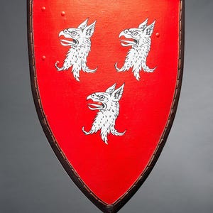 May include: A red shield with a brown leather border and three white griffin heads. The shield has a pointed bottom and is likely a historical or decorative item.