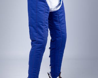 May include: Blue padded fencing pants with red stitching and white accents. The pants have a white crotch protector and are secured with black straps and red ties. Brown leather boots complete the look. The Steel Mastery logo is visible.