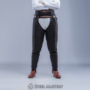 May include: Black quilted padded pants with a brown leather belt and buckles. The pants are worn by a person standing on a gray background.