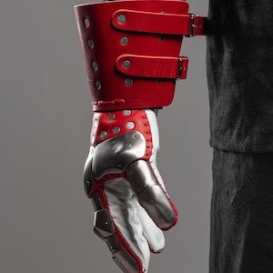 May include: A red leather gauntlet with silver metal plates and white leather. The gauntlet features a red leather cuff with two buckle straps. The hand portion has silver metal plates and red stitching.
