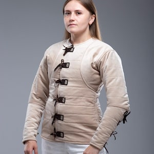 May include: A beige padded gambeson with long sleeves and brown leather ties. The gambeson features a quilted design and is worn over white trousers. The Steel Mastery logo is visible.