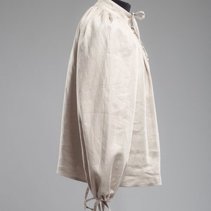May include: A beige linen shirt with long sleeves and a gathered neckline. The sleeves have a gathered cuff with a tie closure.