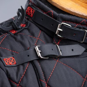 May include: Close-up of a black quilted garment with red stitching. The garment has a leather strap with a silver buckle. The strap is fastened around the neck of the garment. The text "STEEL MASTERY" is visible at the bottom of the image.