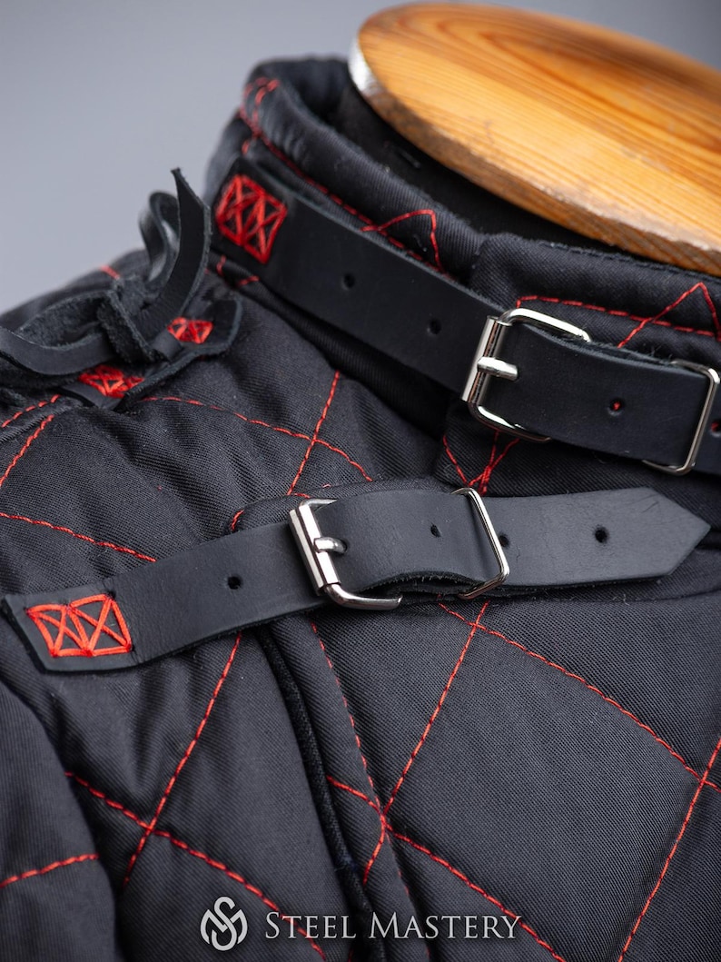 May include: Close-up of a black quilted garment with red stitching. The garment has a leather strap with a silver buckle. The strap is fastened around the neck of the garment. The text "STEEL MASTERY" is visible at the bottom of the image.