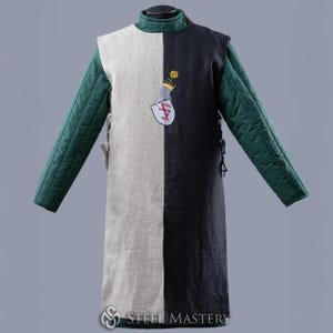 Puede incluir: Gambesón medieval bicolor, dividido verticalmente con un diseño crema y negro. Las mangas son verdes acolchadas. Un escudo con una corona y un escudo está en el lado negro. Diseñado para recreaciones históricas o disfraces.