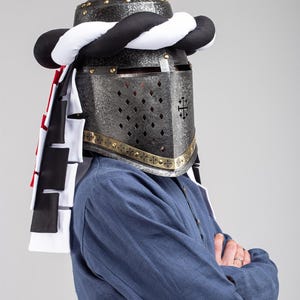 Op de afbeelding: Een zilveren helm met een zwarte en witte turban-achtige hoofdbedekking. De helm heeft een gezichtsvizier met een kruispatroon en is versierd met gouden accenten. De helm wordt gedragen door een persoon die een blauw shirt draagt.