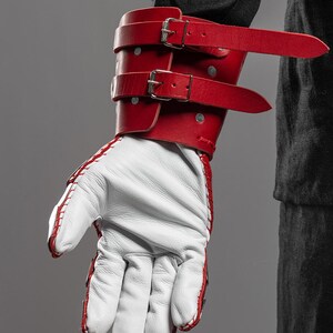 May include: A red leather wrist cuff with two silver buckles and straps, paired with a white leather glove with red stitching. The cuff is secured with silver rivets. The glove is visible with the fingers extended.
