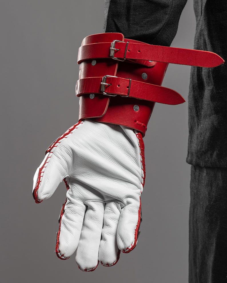 May include: A red leather wrist cuff with two silver buckles and straps, paired with a white leather glove with red stitching. The cuff is secured with silver rivets. The glove is visible with the fingers extended.